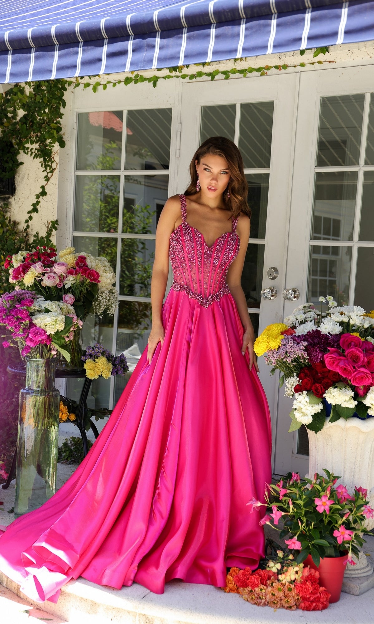 Wearing the Long Formal Dress 29521 by Ava Presley, a woman stands on a porch surrounded by colorful flower arrangements in vases and pots.